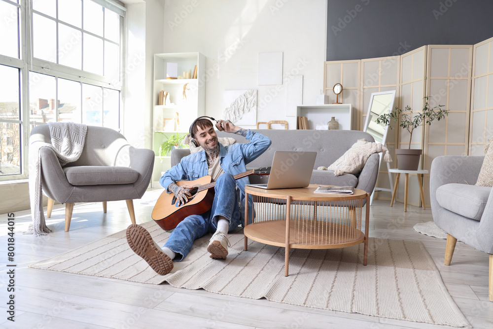 Poster Young tattooed man in headphones playing guitar at home