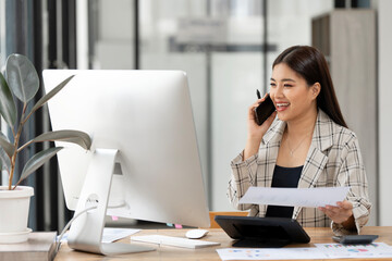 Image of young beautiful joyful woman smiling while using mobile phone and working with desktop computer in office