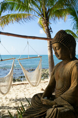 Buddha statue on tropical beach facing the sea, palm trees and hammocks in the background, ideal place for meditation and relaxation 