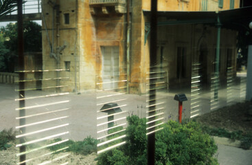 horizontal image of an old yellow building reflected in a glass window. patterns showing horizontal blinds. sunlight and green plants. location Birchircara Rail Station Malta 2003