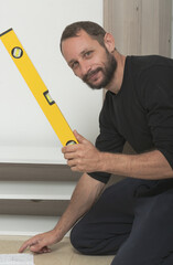 Man kneeling on the floor, holding a yellow building level against a wooden surface, wearing black clothes. Instructions are visible on the floor. Happy smiling male workman using a yellow level tool