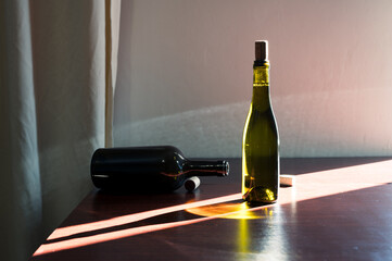 Green glass bottle of wine with cork and opened dark bottle on table in sun ray. Decor winery cellar bar interior photo
