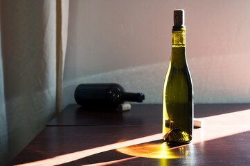 Green glass bottle of wine with cork and opened dark bottle on table in sun ray. Expensive decor winery cellar bar italian france interior