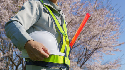 春の桜を背景に誘導棒とヘルメットを持つ作業服の男性｜建設業・警備業などのイメージ