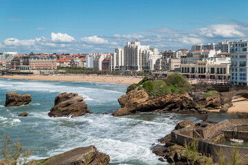 Alguns rochedos no mar junto à praia em Biarritz com parte da cidade ao fundo num dia ensolarado - obrazy, fototapety, plakaty