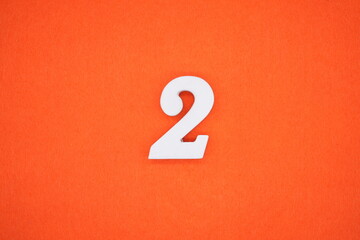 The number 2 is made from white painted wood placed on a background of orange paper.