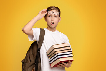 Shocked schoolboy in glasses holding stack of books, orange panorama background with empty space