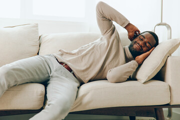 Relaxed African American Man Sitting on a Comfortable Sofa in a Modern Living Room, Smiling and Holding a Smartphone He enjoys his weekend at home, feeling happy and content The background features a
