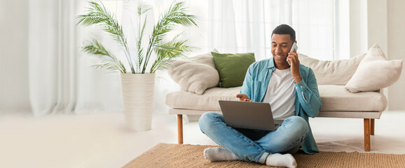 Relaxed African American guy using laptop and chatting on a phone at home, web-banner format with...