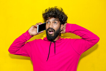 A young Indian man talking on the phone in a pink hoodie calling to discuss plans, looking away with a shocked facial expression. Indoor studio shot isolated on a yellow background