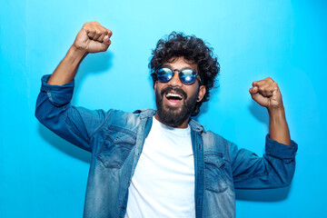 Overjoyed happy Indian curly man screaming yes in ecstatic, raising fists up, celebrating good luck, victory, isolated on Blue. Goal achievement concept