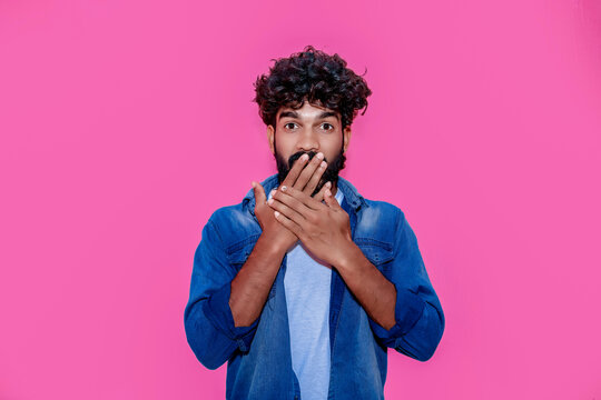 Shocked surprised young indian man looking at camera with omg face expression covering mouth with hands feeling amazed speachless silent reaction standing isolated on pink background.