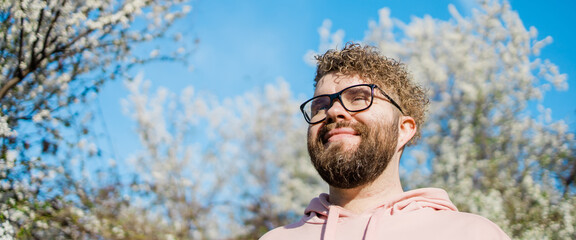 Male bearded man standing under branches with flowers of blooming almond or cherry tree in spring...