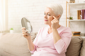 Elderly woman is seated on a couch, her gaze fixed on a mirror in front of her. She appears focused...