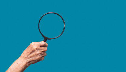 Close-up of an old woman's hand holding a magnifying glass against a blue background.