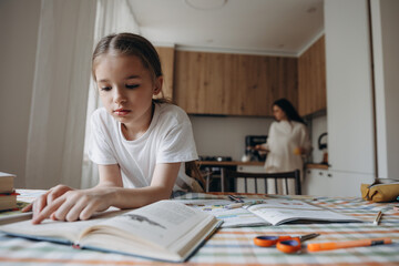 A young girl focused on reading book at a table, with a pregnant mother in the background in a modern kitchen.