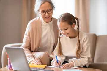 A senior woman with glasses is helping a young girl, who is focused on writing in her notebook, with her homework at a table. They are using a laptop