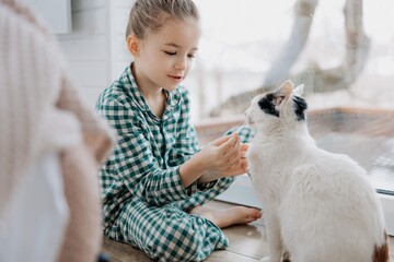 A little blond girl wearing pajamas hugs a domestic cat in her bedroom. The concept of a cozy home morning
