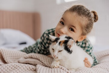 A little blond girl wearing pajamas hugs a domestic cat in her bedroom. The concept of a cozy home...