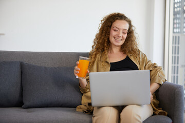 young woman working on laptop computer and holding fresh orange juice at home