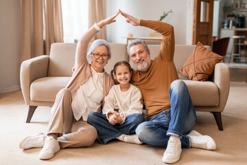 A smiling elderly couple and a young girl sit on the floor in a cozy living room, forming a house...