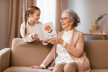 A young girl with a plaited hairstyle is showing a colorful handmade card to her delighted elderly...