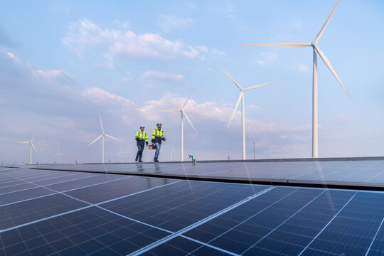 Engineers work walking on factory roof inspect survey and check solar cell panel by hold boxed with turbine wind mill field background is ecology energy alternative power factory concept.