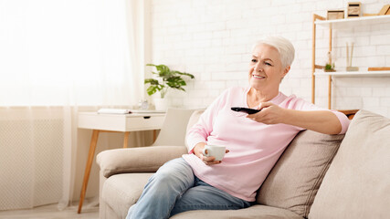 Elderly woman sitting comfortably on a couch, holding a remote control in her hand. She appears to...