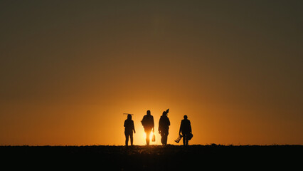 A family of farmers with equipment walks through the field. Silhouettes at sunset. Back view