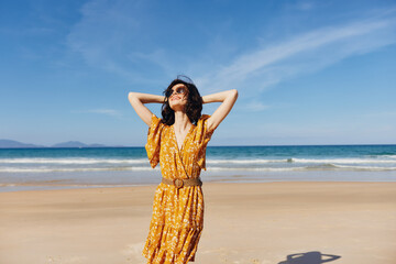 Stunning woman in a vibrant yellow dress posing gracefully on the sandy beach with hands on head