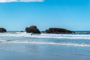 Praia com alguns rochedos e surfistas a desfrutarem das ondas sob um sol radiante no horizonte - obrazy, fototapety, plakaty