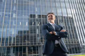 Smiling confident latin hispanic mature business man standing crossed arms. Proud portrait of older senior businessman professional ceo, manager, leader looking aside near office building background