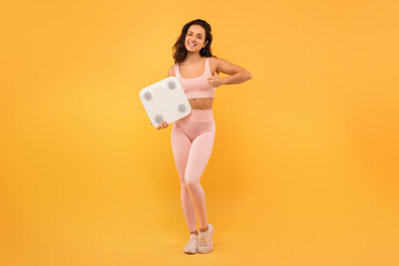 Athletic woman wearing a pink bodysuit is seen holding a scale in her hands. She appears focused...
