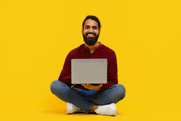 Indian man is seated on the floor, working on a laptop computer. He appears focused and engaged in his task, isolated on yellow background