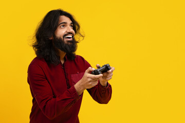 Indian man with a beard and long hair is standing against a bright yellow backdrop, wearing a red...