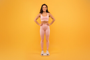 A smiling young woman stands confidently with her hands on her hips, dressed in a light pink sports bra and leggings, checking her weight