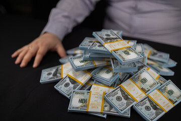 Side view of a large pile of stacks of dollars on a black background next to a seated man. The man...