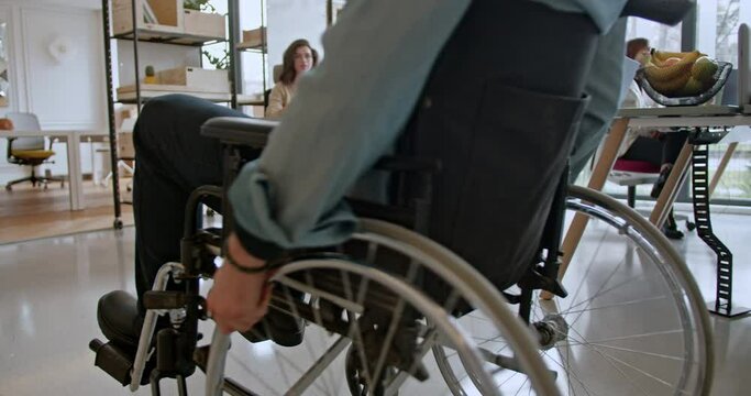 In a multicultural and inclusive office setting, businessperson in a wheelchair rolls towards a woman colleague working at a desk, highlighting accessibility and equal participation in the workplace.