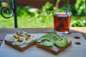 Open face sandwiches spread by fresh cheese and garnished with hard boil egg and slices of riped avocado, served outside in the sunny garden as a healthy and balanced breakfast with cup of black tea.