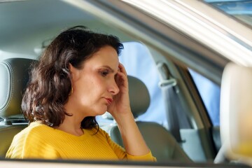 Worried woman in vehicle, hand on forehead, contemplative gaze to side, car interior background, natural light, emotion of concern. Service work in car repair shop
