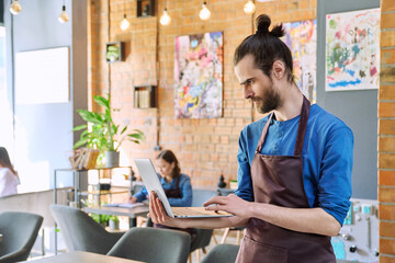 Man service worker owner in apron using laptop in restaurant cafeteria interior