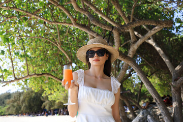 An Asian woman in a casual suit relaxes at the beach beside the sea, holding a glass of orange juice and raising her hand in happiness.