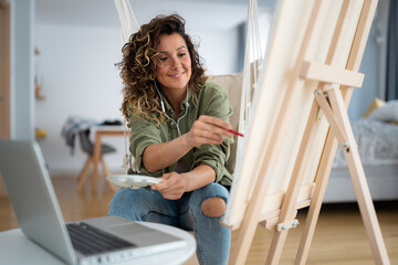 Happy Caucasian woman smiling while painting on a canvas at home and listening to music or podcast.