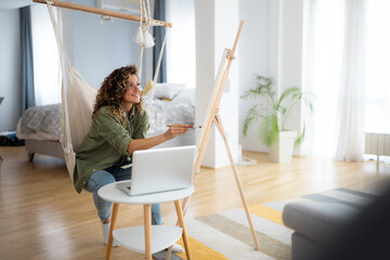 Beautiful young woman artist painting in a living room at home and also using a laptop for her work.
