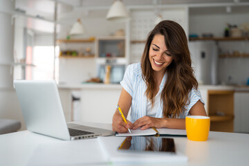 Smiling young woman using laptop sitting at the table in a home office. Focused female entrepreneur...