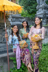 Three young women in traditional Balinese attire, holding offerings and a ceremonial umbrella,...