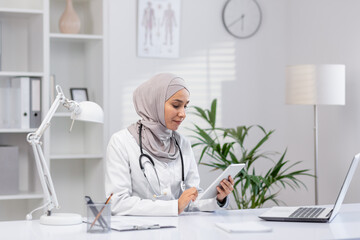 A professional Muslim female doctor in a hijab sits in her office, using a digital tablet. The...