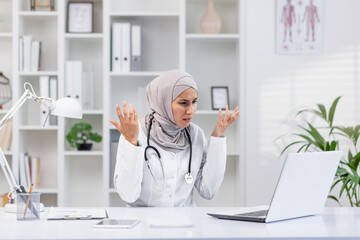 Professional female doctor wearing a hijab looks frustrated while working on her laptop in a modern...