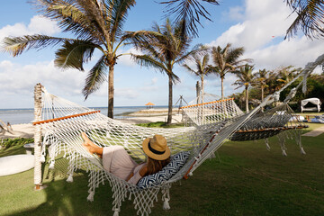 Elegant woman chilling  in a hammock strung between palm trees on the beach at a tropical resort,...