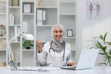 A cheerful female doctor wearing a hijab greets with a friendly wave while sitting at her desk in a...